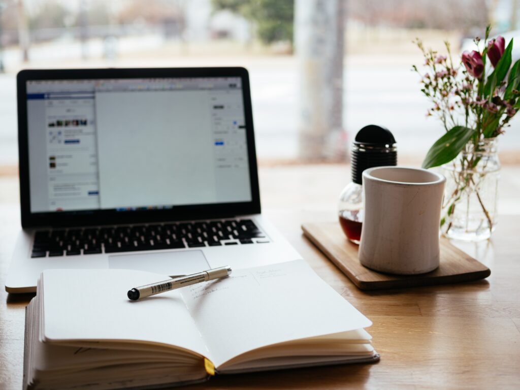 Notebook with pen along with laptop open with screen on and coffee mug on the left.